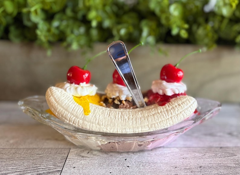 Pu&ograve; includere: Un banana split sundae con tre palle di gelato, guarnito con panna montata, ciliegie e un cucchiaio. Il banana split &egrave; in una ciotola di vetro.