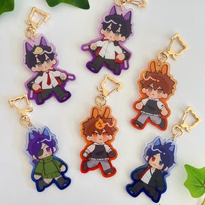 May include: Six colorful character keychains with gold clasps. Each keychain features a different character with a unique design and color scheme. The characters are all wearing different outfits and have different expressions.