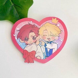 May include: A heart-shaped sticker with two cartoon characters. The character on the left has red hair and is wearing a white shirt with a red bow. The character on the right has blonde hair and is wearing a blue and white outfit with a crown. The sticker has a pink and blue background with white hearts.