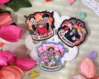 May include: Three decorative enamel pins featuring illustrated characters. One pin shows three figures with the text THE SPANNERS, another shows three figures with THE BAD BOYS, and the third shows three figures with the text BAMBOOZLERS.