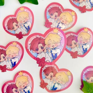 May include: A set of 10 heart-shaped stickers featuring two cartoon characters. The characters are a blonde-haired person with a crown and a red-haired person. The stickers are pink and white with a glittery finish.