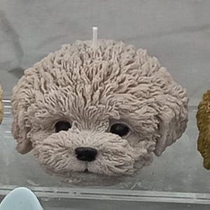May include: Three dog-shaped candles in a row. The candles are in shades of cream, grey, and brown. Each candle has a detailed face with a black nose and eyes, and a white wick. The candles are on a clear surface.