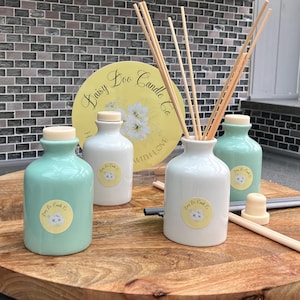 May include: Four ceramic diffuser bottles with a white and yellow label that says "Daisy Doo Candle Co. Handmade with Love". Two bottles are light green and two bottles are white. The bottles are on a wooden table with a set of wooden diffuser sticks.