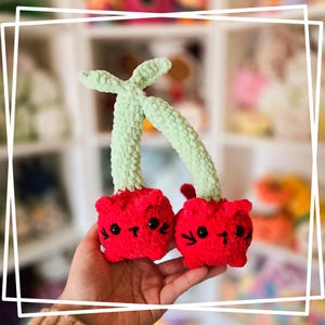 May include: Two crocheted cherry plush toys with green stems and red bodies. The cherries have cute cat faces with black eyes and whiskers.