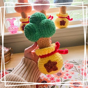 May include: A crocheted plush toy shaped like a money tree. The tree is green and has a brown trunk. The pot is yellow with a red ribbon and a brown star.