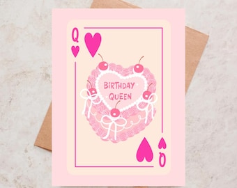 May include: A pink greeting card featuring a heart-shaped cake with cherries and the words "BIRTHDAY QUEEN." The card is designed as a playing card, with the Queen of Hearts suit symbols. The card is on a brown envelope.