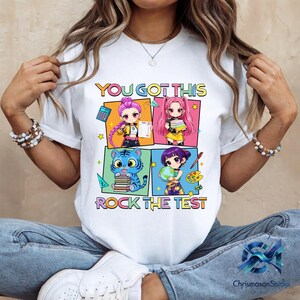 May include: White t-shirt with a colorful graphic that says "YOU GOT THIS ROCK THE TEST". The graphic features four anime-style characters and a tiger holding books. The shirt is worn by a person.