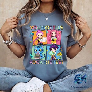 May include: A blue t-shirt with the text "YOU GOT THIS ROCK THE TEST" and a colorful graphic featuring four anime-style characters. The graphic includes a calculator, books, a globe, and paint supplies.