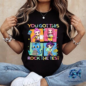 May include: Black t-shirt with the text "YOU GOT THIS ROCK THE TEST" and colorful anime-style illustrations. The illustrations feature four different characters with books, a calculator, and a globe.