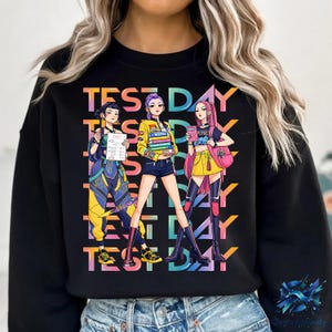 Könnte beinhalten: Schwarzes Sweatshirt mit einer farbenfrohen Grafik, die drei Figuren im Anime-Stil zeigt, die testbezogene Gegenstände halten. Die Worte "TEST DAY" sind in einer lebendigen, regenbogenartigen Schriftart vertikal über dem Design angeordnet. Die Figuren sind in stilvollen Outfits gekleidet.