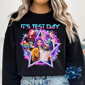 Könnte beinhalten: Schwarzes Sweatshirt mit dem Text "IT'S TEST DAY" in einer farbenfrohen Grafik. Das Design zeigt drei Mädchen im Anime-Stil, einen Stapel Bücher und einen Testbogen mit der Note A, vor einem sternförmigen Hintergrund.