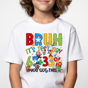 May include: White t-shirt with a colourful graphic that reads "BRUH It's Test Day 67+33% YOU GOT THIS." The design features superhero characters and a playful font. The shirt is made of a soft material.