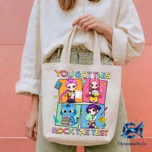 May include: A white tote bag with the text "YOU GOT THIS" and "ROCK THE TEST" in colorful letters. The bag features four cartoon images of anime-style characters. The bag is held by a person wearing a white shirt.