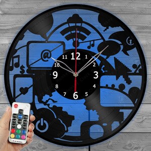 May include: A blue and black vinyl record clock with a remote control. The clock face features a design of various internet and technology icons, including a laptop, a wifi symbol, a music note, a dollar sign, a heart, a power button, and a camera. The clock face is surrounded by a black vinyl record with a white border. The clock has a black metal frame and a black metal hand.