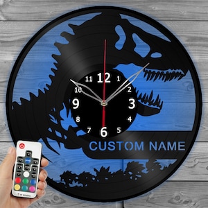 Puede incluir: Un reloj de vinilo negro con un fondo azul que presenta la silueta de un dinosaurio. La esfera del reloj tiene números y manecillas negros. El reloj tiene un control remoto con múltiples luces de colores. El texto "CUSTOM NAME" está impreso en la esfera del reloj.
