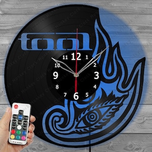 May include: A black vinyl record clock with a blue flame design and the word "TOOL". The clock has a black face with white numbers and hands. A remote control is held in the lower left corner.