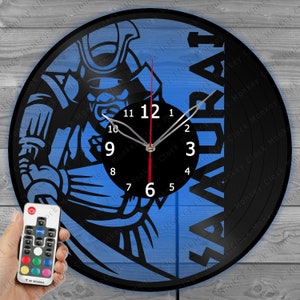 Op de afbeelding: Een zwarte vinyl platenspeler klok met een blauwe en zwarte afbeelding van een samoerai-krijger. De wijzerplaat van de klok is wit met zwarte cijfers en wijzers. De klok is verlicht met LED-verlichting en wordt geleverd met een afstandsbediening.