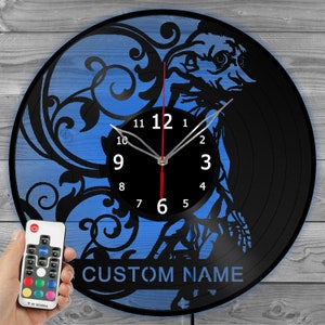 Könnte beinhalten: Eine schwarze Schallplattenuhr mit blauem Hintergrund und dekorativem Design. Das Zifferblatt ist schwarz mit weißen Zahlen und Zeigern. Der Text "CUSTOM NAME" steht unten. Eine Fernbedienung ist im Bild.