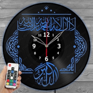 Może przedstawiać: Czarny zegar winylowy z niebieskim wzorem arabskiej kaligrafii i czerwoną i srebrną tarczą. Zegar jest podświetlany diodami LED i wyposażony w pilot.
