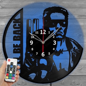 May include: Black vinyl record clock with a silhouette of a person against a blue background. The clock face is black with white numbers and hands. The text "BE BACK" is on the left side. A remote control is in the lower left corner.