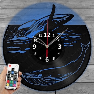 May include: A black vinyl record clock with a blue whale design. The clock has a white face with black numbers and hands. The clock is illuminated with a blue light. The clock comes with a remote control to change the color of the light.