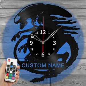 May include: Black vinyl record clock with a blue and gray background. The clock features a silhouette of an alien creature and the text "CUSTOM NAME". The clock hands are silver, and the second hand is red. A remote control is in the image.
