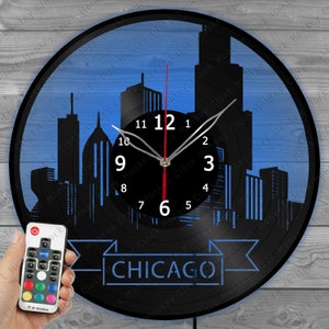 Könnte beinhalten: Eine schwarze Schallplattenuhr mit blauem Hintergrund, die eine Silhouette der Skyline von Chicago zeigt. Das Zifferblatt der Uhr ist weiß mit schwarzen Zahlen und Zeigern. Die Uhr verfügt über eine Fernbedienung mit farbwechselnden LED-Lichtern. Der Text "CHICAGO" wird in einem blauen Banner am unteren Rand der Uhr angezeigt.