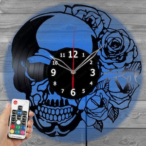 May include: A black vinyl record clock with a skull and roses design. The clock face is white with black numbers and hands. The clock is set against a blue background. The clock is illuminated with colorful LED lights that can be controlled with a remote.