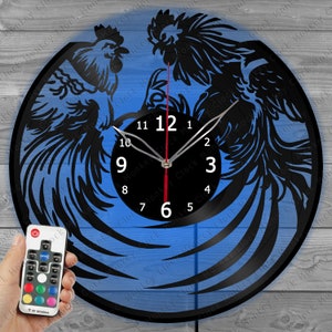 Könnte beinhalten: Schwarze Hahn-Silhouette-Uhr mit blauem Hintergrund. Die Uhr hat ein schwarzes Zifferblatt mit weißen Zahlen und silbernen Zeigern. Eine Fernbedienung befindet sich in der unteren linken Ecke. Die Uhr besteht aus einer Schallplatte.