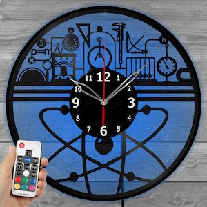 May include: A blue and black vinyl record clock with a science theme. The clock features a stylized atom, a beaker, a compass, and other science-related imagery. The clock has a black frame and a white clock face with black numbers. The clock is illuminated with a blue light.