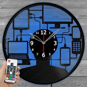 May include: A black vinyl record clock featuring a silhouette of a person's head at its centre. The clock face is surrounded by a design of various electronic devices, including a laptop, a desktop computer, a smartphone, and a tablet. The clock has a red second hand and a white minute and hour hand. The clock is illuminated with a blue light. The clock comes with a remote control with multiple colour options.