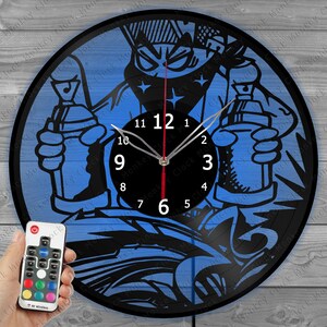 May include: A black vinyl record clock with a blue and black graphic of a person holding spray paint cans. The clock has a black face with white numbers and hands. The clock is illuminated with colorful LED lights and comes with a remote control.