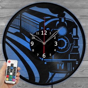 Puede incluir: Un reloj de vinilo negro con una silueta azul y negra de una locomotora. El reloj tiene una esfera blanca con números y manecillas negros. El reloj está iluminado con una luz azul. Un mando a distancia está en primer plano.