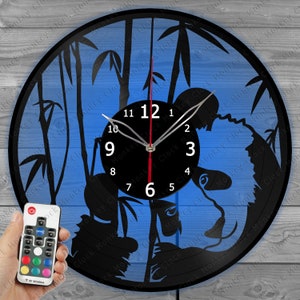 Puede incluir: Un reloj de vinilo negro con un diseño de oso panda. El reloj tiene un fondo azul con tallos de bambú. El reloj está iluminado con luces LED y viene con un control remoto para cambiar el color de las luces.