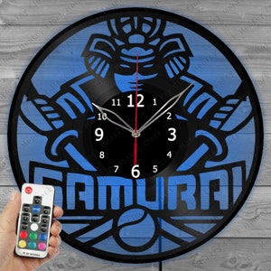 Op de afbeelding: Een blauwe en zwarte vinyl platenspeler klok met een samurai-krijger ontwerp en het woord "SAMURAI" in witte letters. De klok heeft een zwarte wijzerplaat met witte cijfers en een rode secondewijzer. De klok wordt verlicht met een blauw licht en wordt geleverd met een afstandsbediening.