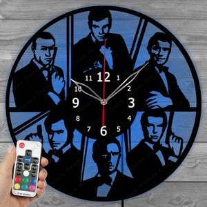 Puede incluir: Un reloj de pared negro con siluetas de varias figuras sobre un fondo azul. El reloj tiene un diseño circular con un segundero rojo y números blancos. Un mando a distancia es visible en la esquina inferior izquierda.