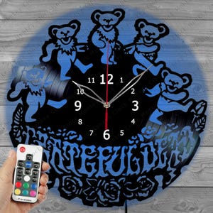 May include: Black and blue wall clock featuring dancing bear silhouettes and the word "Grateful" at the bottom. The clock has a black face with white numbers and red hands. A remote control is held in the lower left corner.