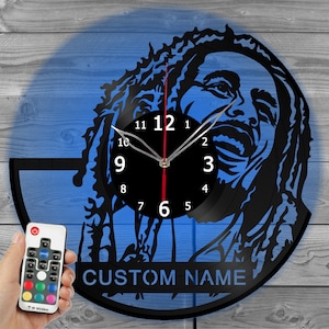 May include: Black wall clock featuring a portrait design. The clock has a black face with white numerals and hands. The clock's design includes the text "CUSTOM NAME" and comes with a remote control.