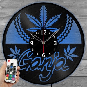 Op de afbeelding: Een zwarte vinyl platenspeler klok met een blauw marihuanablad ontwerp en het woord "Ganja" in blauwe letters. De klok heeft een witte wijzerplaat met zwarte cijfers en wijzers. De klok wordt verlicht met een blauw licht en wordt geleverd met een afstandsbediening.