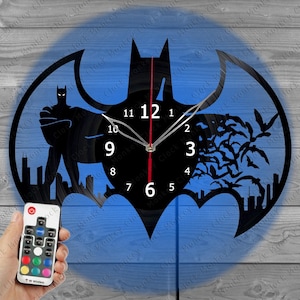 Pode incluir: Relógio de parede com o logotipo do Batman em preto com fundo azul. O relógio apresenta uma silhueta do Batman, silhuetas de cidades e morcegos. Os ponteiros do relógio são prateados com um ponteiro de segundos vermelho. Um controle remoto está na imagem.