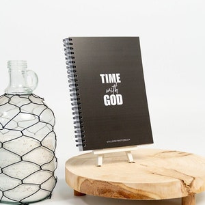 Könnte beinhalten: Ein schwarzes spiralgebundenes Notizbuch mit dem Text "TIME with GOD" und den Worten "Stillzeitnotizbuch" in kleinerer Schrift. Das Notizbuch steht auf einem Holzständer und eine klare Glasflasche mit einem Drahtgeflecht befindet sich im Vordergrund.