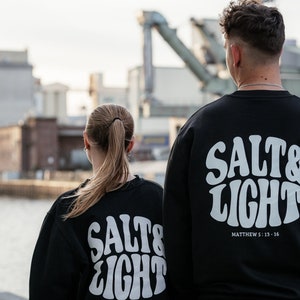 Könnte beinhalten: Zwei Personen tragen schwarze Sweatshirts mit weißem "SALT & LIGHT"-Text auf der Rückseite. Der Text ist in einem Retro-Schriftart-Stil gehalten. Der Text enthält auch "Matthew 5:13-16" darunter.