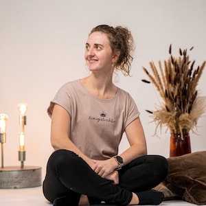 Könnte beinhalten: Eine Frau sitzt im Schneidersitz auf einer weißen Oberfläche und trägt ein hellbraunes T-Shirt mit dem Text "königstochter" und einer Krone über dem Text. Sie trägt eine schwarze Hose und schwarze Socken.