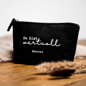 Könnte beinhalten: Eine schwarze Reißverschlusstasche mit den Worten "Du bist wertvoll" in weißer Schrift. Der Text "JESAJA 43,4" steht darunter. Die Tasche liegt auf einer Holzoberfläche mit einem flauschigen, hellfarbigen Material.