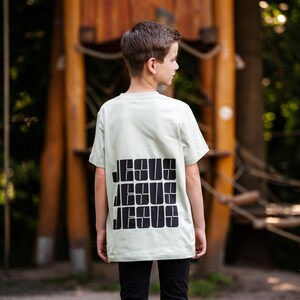Könnte beinhalten: Ein junges Kind trägt ein hellgrünes T-Shirt mit dem Wort "JESUS" in schwarzen Blockbuchstaben auf dem Rücken. Das Kind steht vor einer Holzkonstruktion.