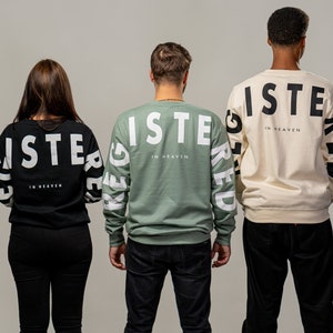 Könnte beinhalten: Drei Personen tragen schwarze, grüne und cremefarbene Sweatshirts mit dem Wort "REGISTERED" auf dem Rücken, das rückwärts geschrieben ist. Der Text "IN HEAVEN" ist in kleineren Buchstaben unter dem Wort "REGISTERED" gedruckt.