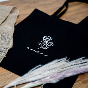 Könnte beinhalten: Schwarze Einkaufstasche mit einem weißen Blumenmuster und dem Wort "amen" in Kursivschrift.