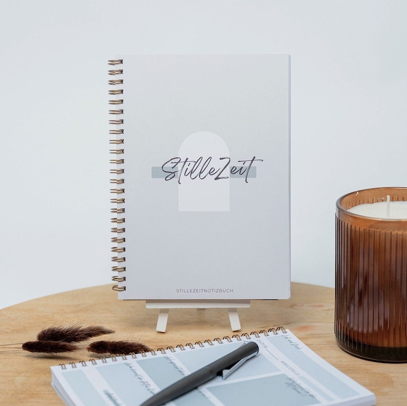 May include: A gray spiral bound journal with the text "StilleZeit" and "StilleZeitNotizbuch" on the cover. The journal is on a white easel and a brown wooden table. A brown candle is in the background.