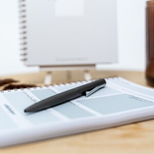 May include: A black pen lying on a white planner with a blue grid pattern. The planner has a brown spiral binding. The planner is on a wooden surface.