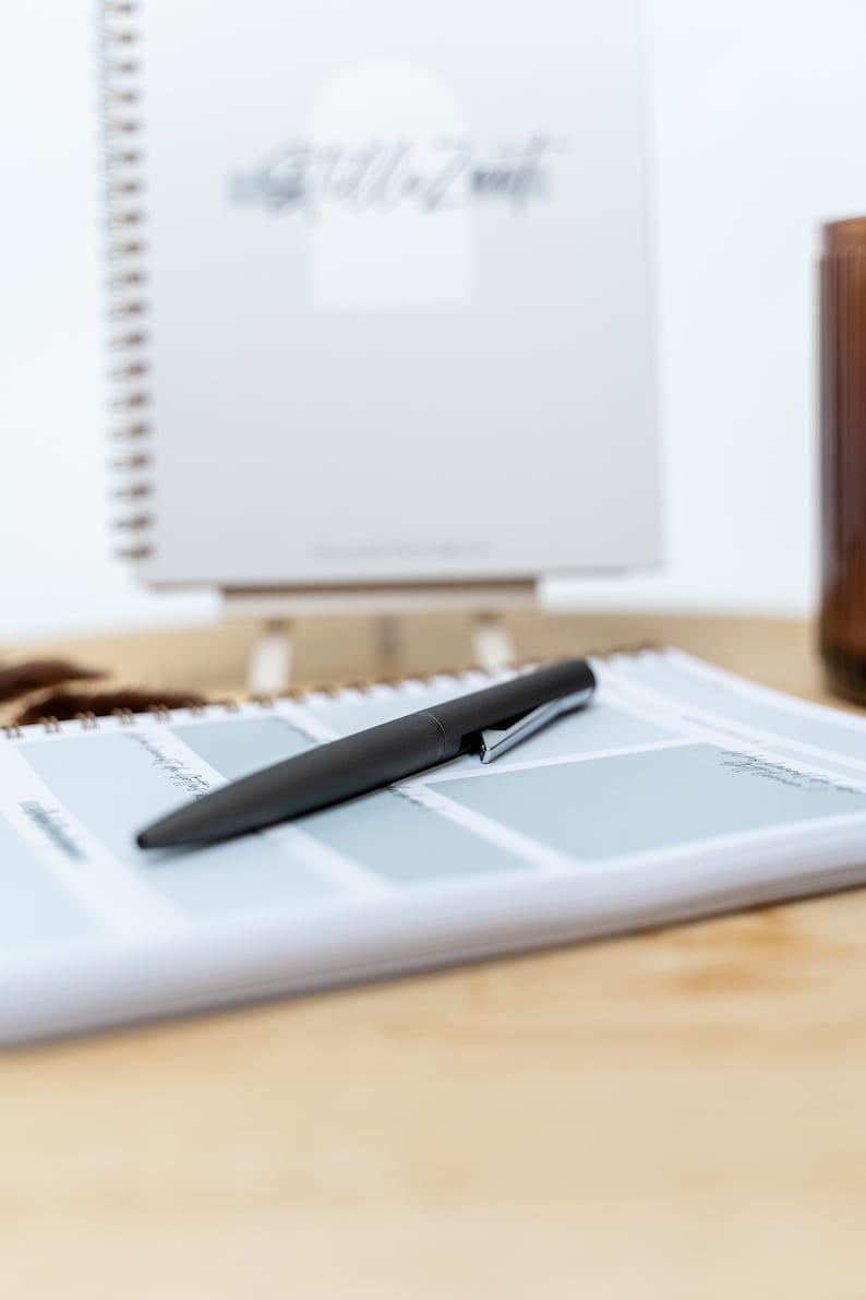 May include: A black pen lying on a white planner with a blue grid pattern. The planner has a brown spiral binding. The planner is on a wooden surface.